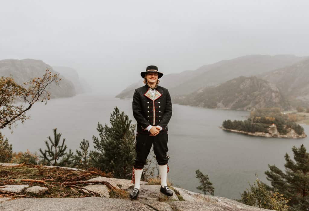 Bryllupsfotografi av en person i tradisjonell drakt med fjell- og fjordlandskap i bakgrunnen.